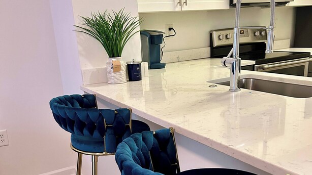 A modern kitchen with white cabinets, a stainless steel sink, and a coffee maker. There are two blue bar stools with gold legs. - Chic Blue Apt w Patio + Pool, Free Parking, 10 min to ATL (Atlanta)