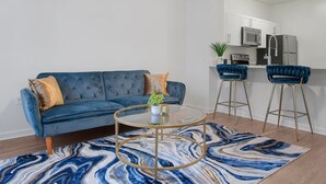 A modern living room with a blue sofa, a glass coffee table, and a kitchen area with a refrigerator and microwave. - Chic Blue Apt w Patio + Pool, Free Parking, 10 min to ATL (Atlanta)