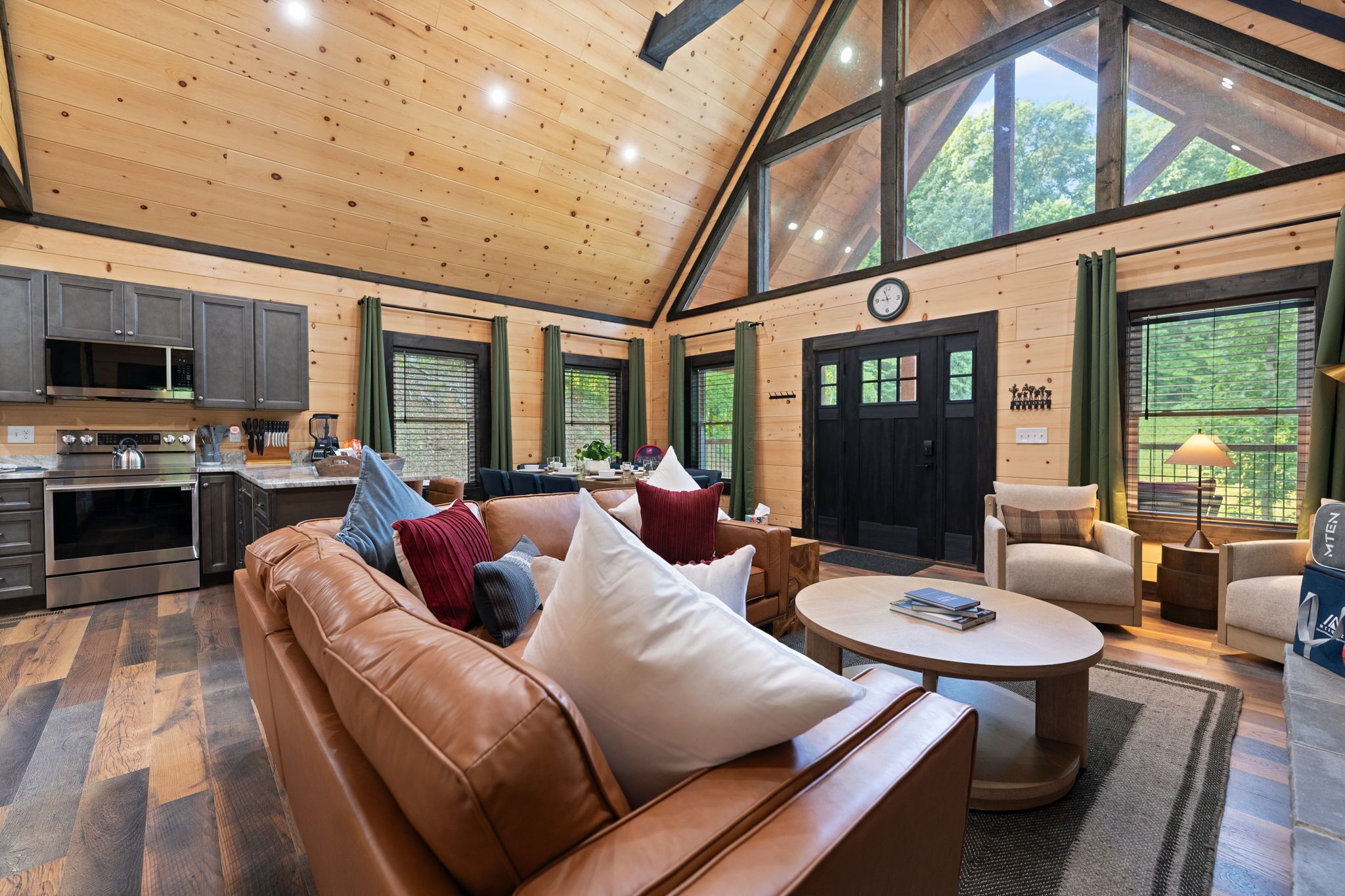 A spacious living area with a leather sofa, a coffee table, and a dining table with chairs. There is a kitchen area with modern appliances and a large window providing a view of the outdoors.