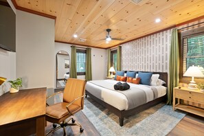 A hotel room with a wooden ceiling, a large bed with multiple pillows, a desk with a chair, and a nightstand with a lamp.