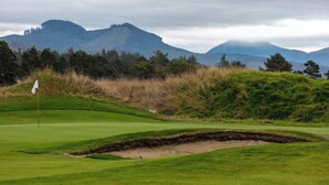 Een golfbaan met vlag, bunkers en een bergachtig achtergrond.