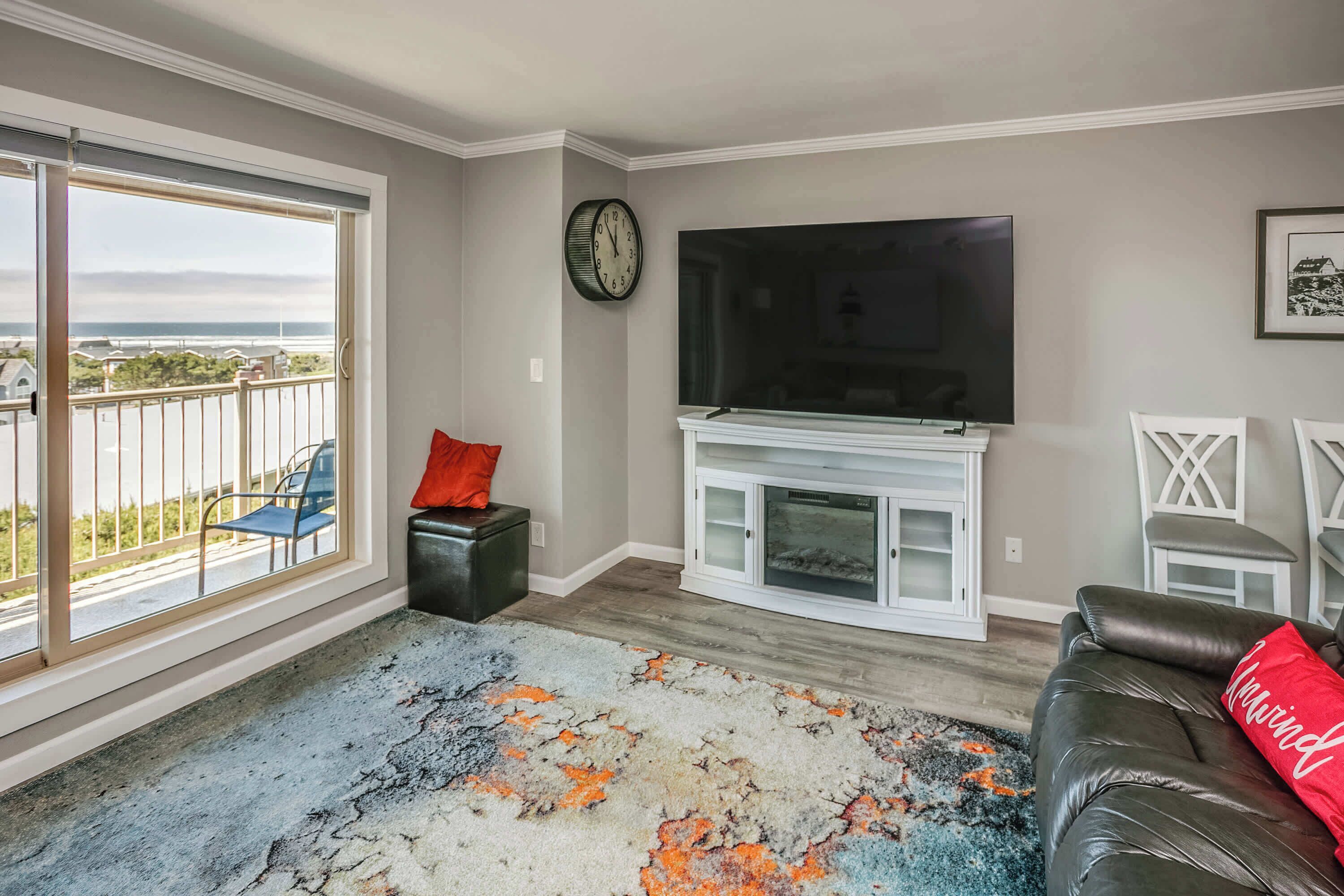 A living room with a large TV, a fireplace, a balcony with a view, and a patterned rug.