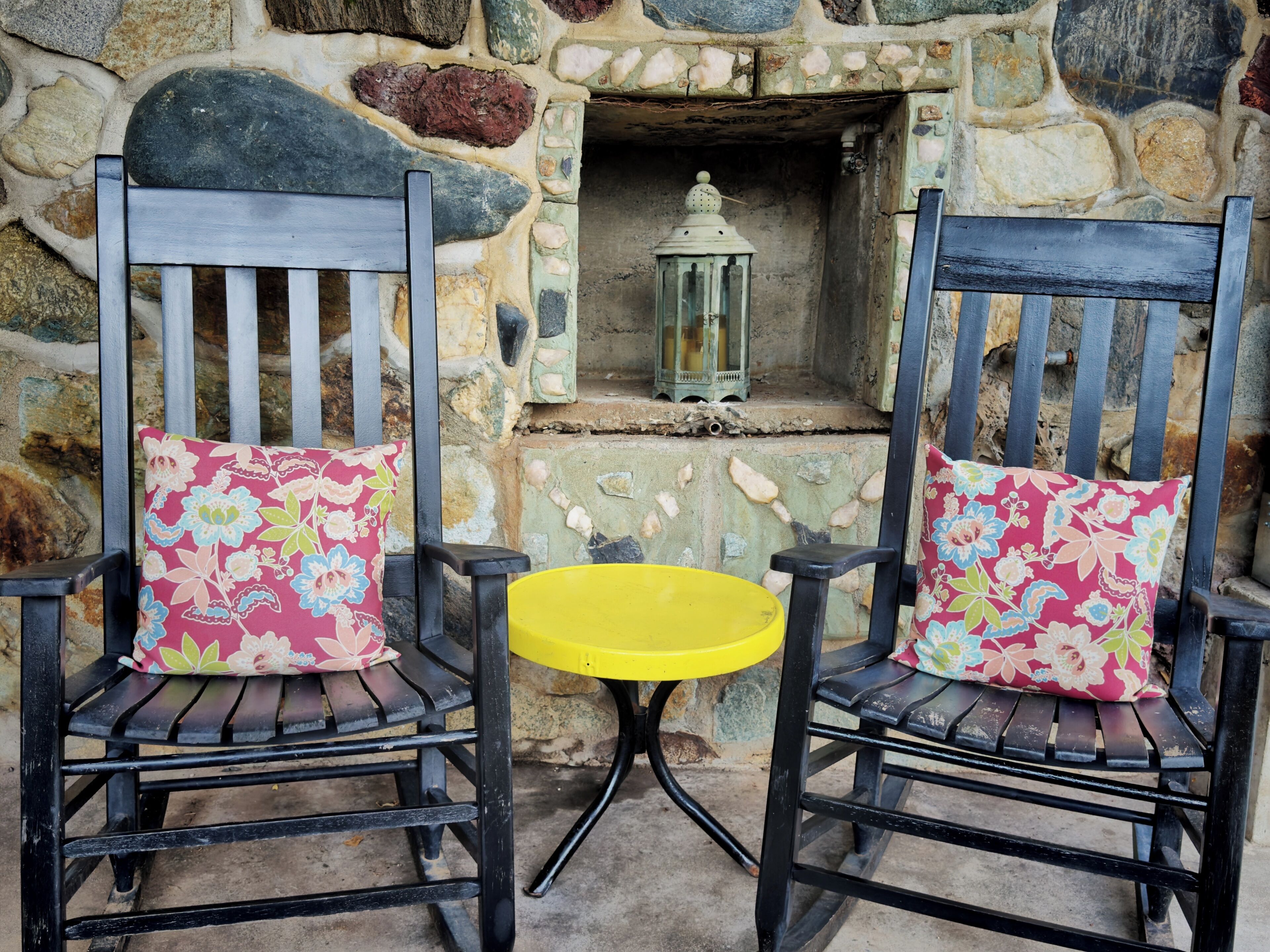 Two black chairs with floral cushions, a small yellow table, and a stone wall with a lantern.