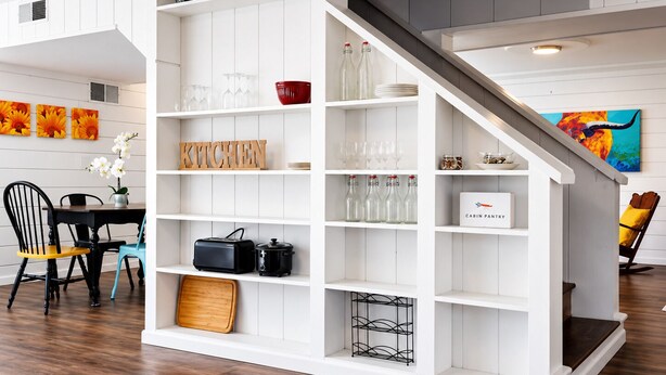 Una cocina moderna con armarios blancos, una zona de comedor con mesa y sillas negras, y una escalera que sube a otro nivel.