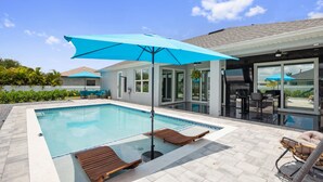 Ein moderner Poolbereich mit Liegestühlen, einem blauen Sonnenschirm und einer verglasten Terrasse.