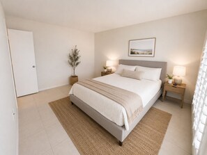 Ein Schlafzimmer mit einem Bett, zwei Nachttischen, einem Beistelltisch, einer Topfpflanze und einem gerahmten Bild an der Wand.