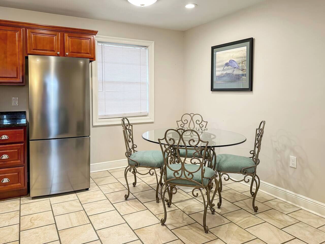 A kitchen with a stainless steel refrigerator, a dining table with four chairs, and a window with blinds.