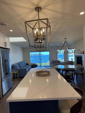 A modern kitchen with a central island, a dining area with a view, and a living area with a sofa. - Coastal Elegance - 2BDRM (Nanoose Bay)