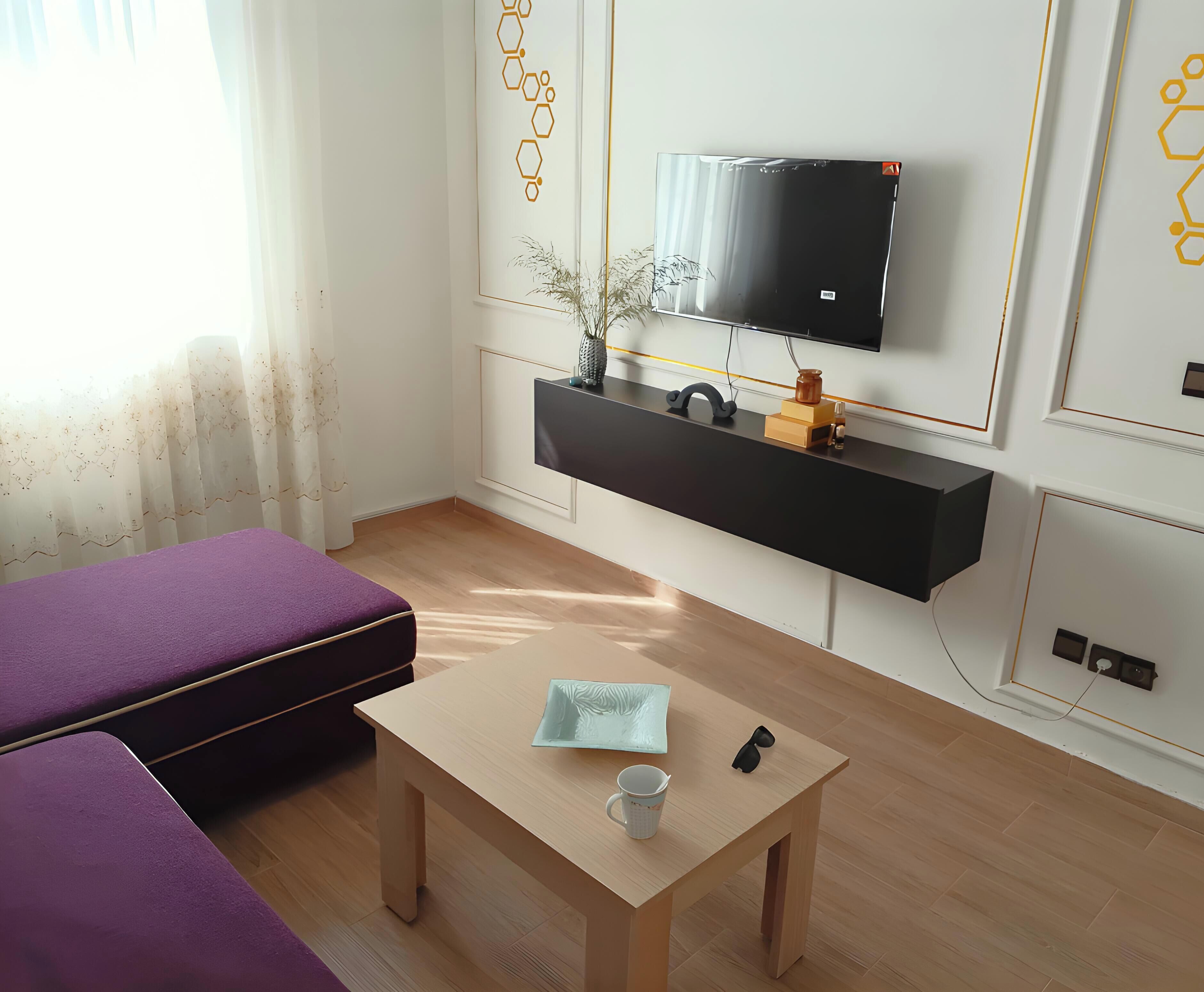 Una camera moderna con un divano, un tavolino con una tazza e occhiali da sole, una TV su un mobile nero e una mensola a parete con oggetti decorativi.