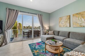 Ein Wohnzimmer mit einer Couch, einem Couchtisch und einem Fernseher. Ein Balkon bietet Blick auf das Meer und Palmen.