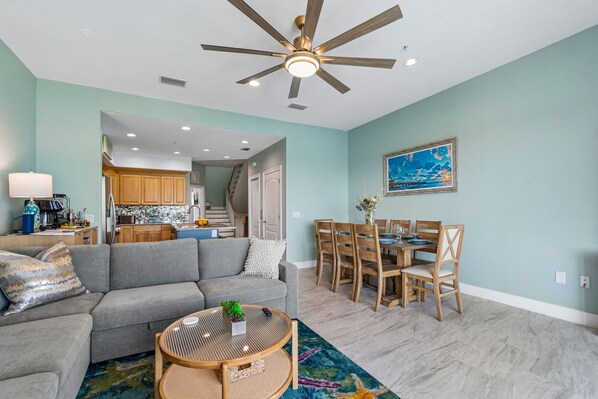 A modern living room with a gray sofa, a dining table with wooden chairs, a ceiling fan, and a kitchen in the background.