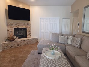 A living room with a fireplace, a flat-screen TV mounted above it, a sectional sofa, and a coffee table with a vase. - Beautifully decorated single-level private townhome in the heart of Oro Valley. (Tucson)