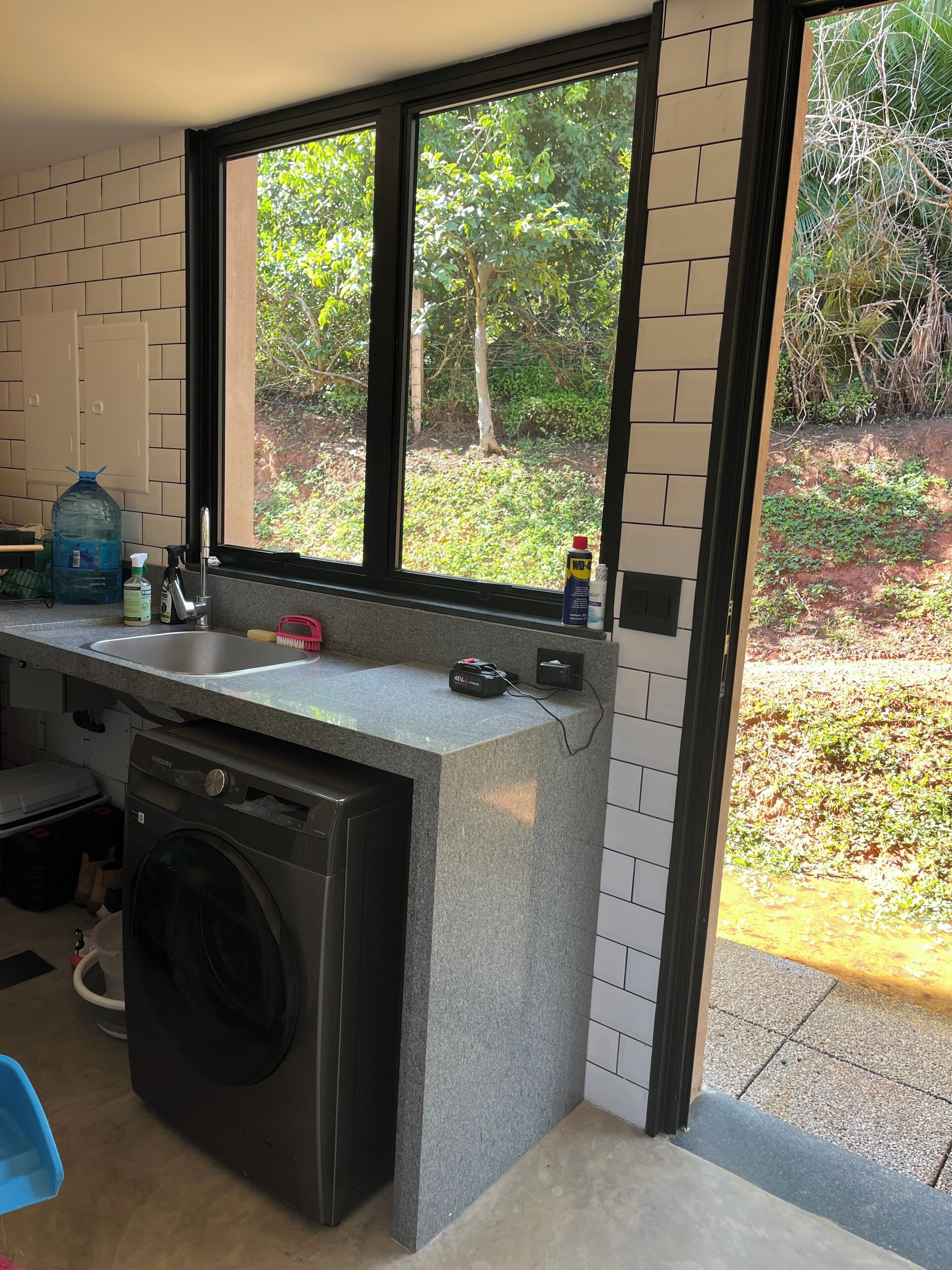 A kitchen with a washing machine, a large water dispenser, and a window with a view of greenery.