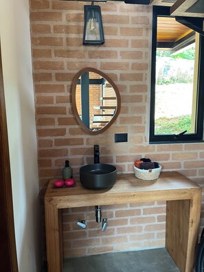 A bathroom with a round mirror, a black basin, a wooden vanity, and a window with a view of greenery.