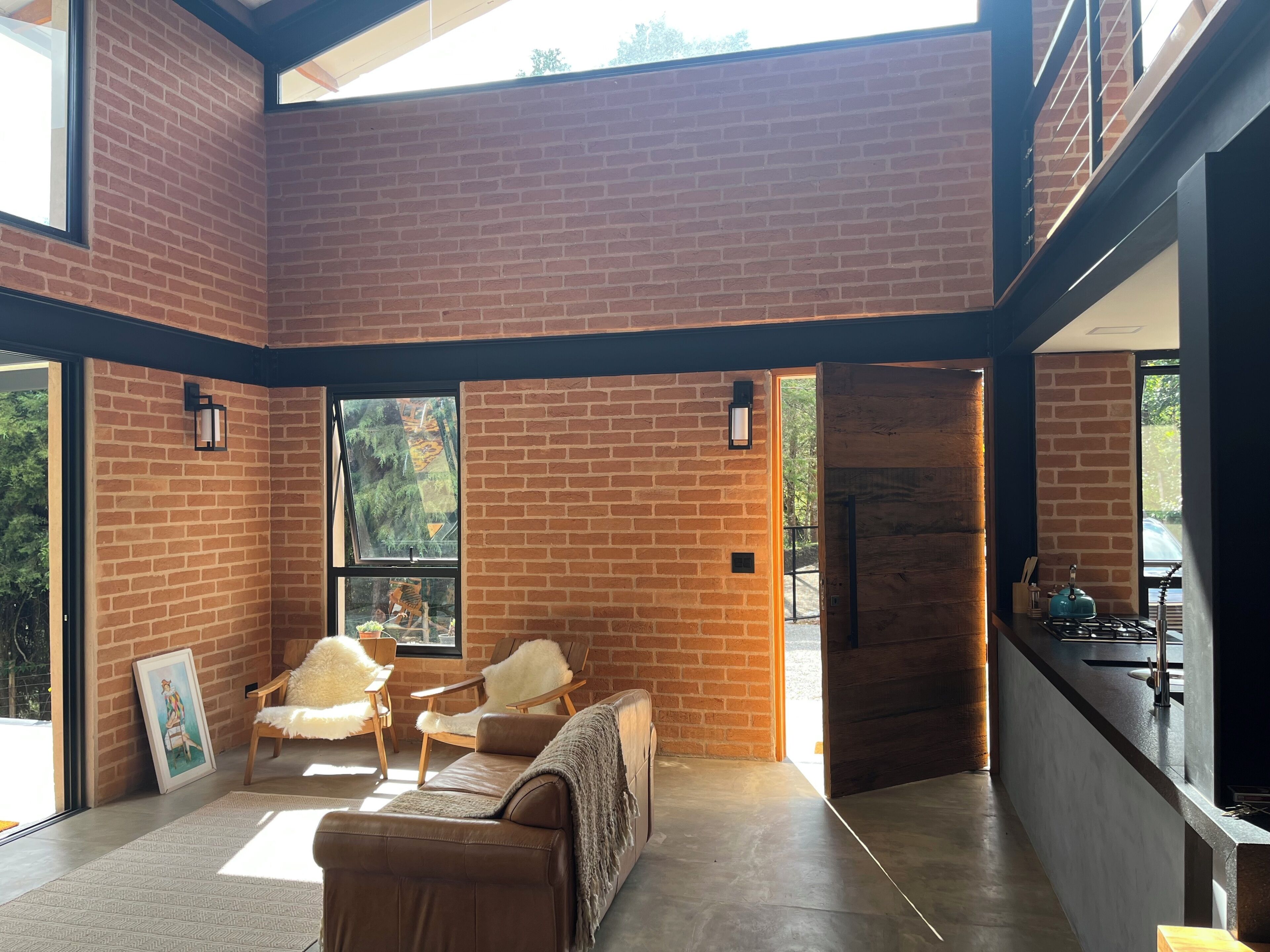 Una habitación amplia con pared de ladrillo, un sofá, dos sillones, un cuadro y una zona de cocina con estufa.