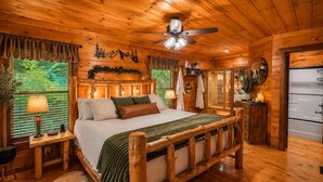 Ein Schlafzimmer in einer Blockhütte mit einem hölzernen Bett, einem Deckenventilator, einem Badezimmer mit Badewanne und einem Fenster mit Jalousien.