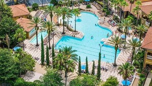 A large outdoor swimming pool surrounded by palm trees and lounge chairs, with a parking area visible in the background. - 3BR Resort Suite on I-drive + Pools (Orlando)