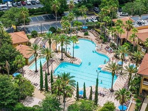 Une grande piscine extérieure entourée de palmiers et de chaises longues, avec un parking visible au loin.