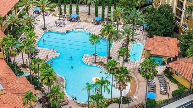 Ein großer Swimmingpool, umgeben von Palmen und Liegestühlen, mit einem Gebäude im Hintergrund.