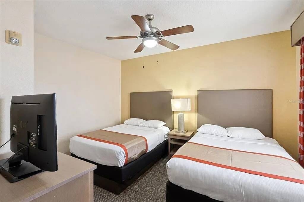 A hotel room with two beds, a ceiling fan, a TV, and a lamp.
