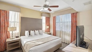 A hotel room with a bed, a desk with a TV, a ceiling fan, and large windows with curtains. - 3BR Resort Suite on I-drive + Pools (Orlando)