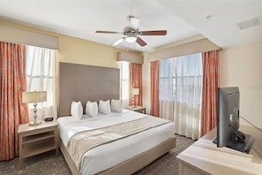 Une chambre d’hôtel avec un lit, un bureau avec une télévision, un ventilateur de plafond et de grandes fenêtres avec des rideaux.
