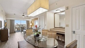 Ein Hotelzimmer mit einem runden Glastisch, Korbstühlen, einer Couch, einem Deckenventilator, einem großen Spiegel und einem Balkon mit Blick.