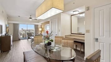 A hotel room with a round glass table, wicker chairs, a sofa, a ceiling fan, a large mirror, and a balcony with a view.