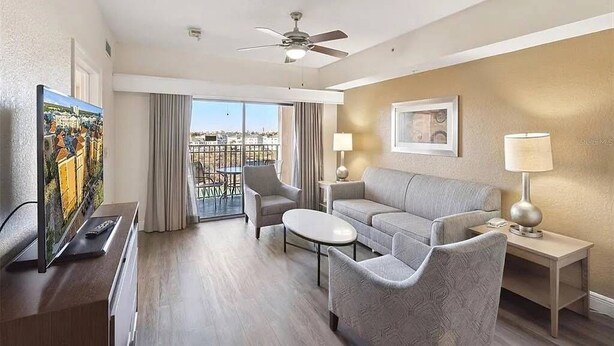 A hotel room with a balcony, a ceiling fan, a sofa, a coffee table, and a TV. - 3BR Resort Suite on I-drive + Pools (Orlando)
