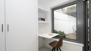 Ein Eckzimmer mit einem weißen Schrank, einem kleinen weißen Regal mit Büchern, einem braunen Sessel und einem Fenster mit Blick auf einen weißen Zaun und Grünflächen.