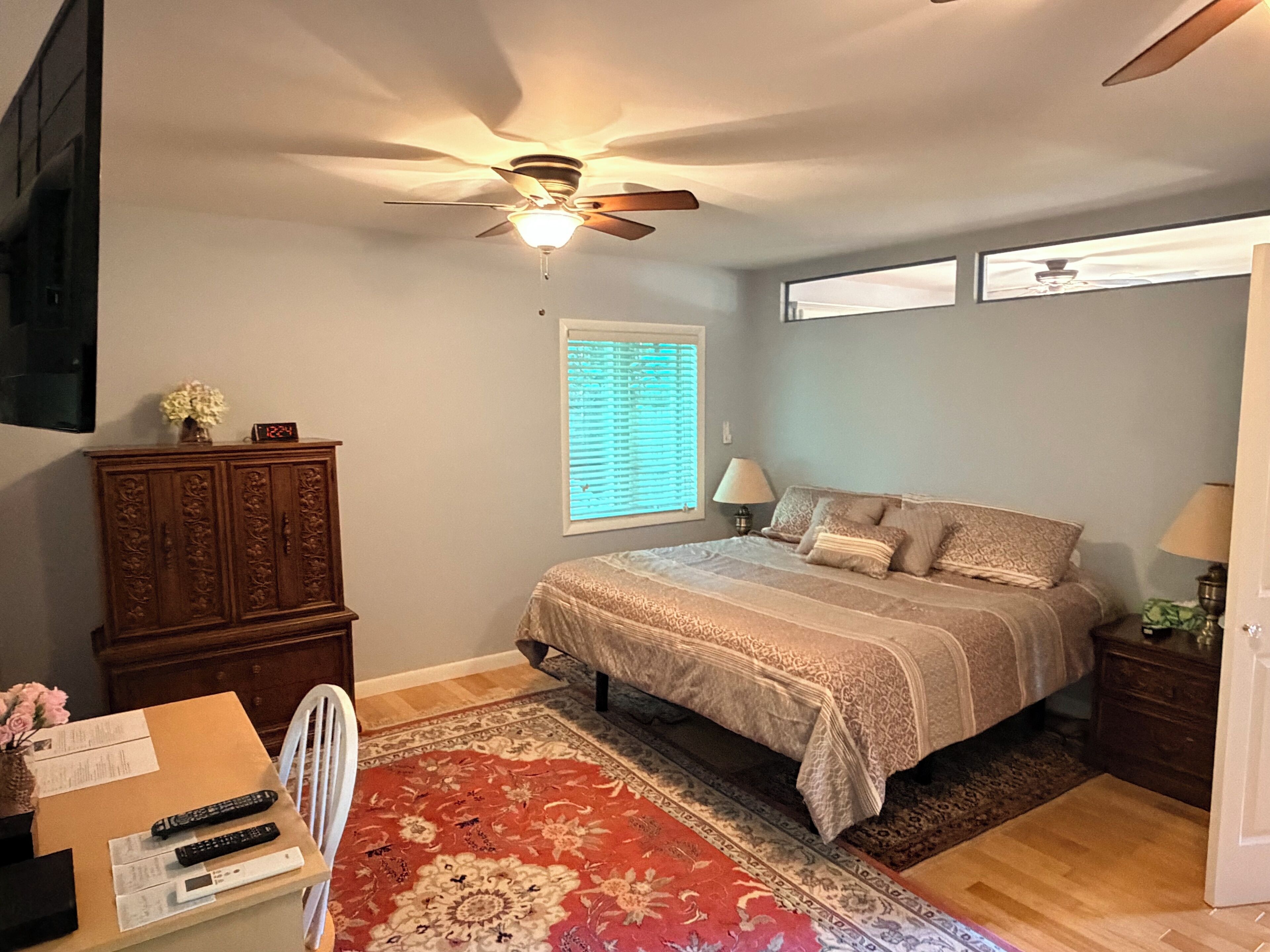 Ein Schlafzimmer mit Bett, Deckenventilator, Fenster mit Jalousien, Nachttisch, Lampe, Kommode und Teppich.