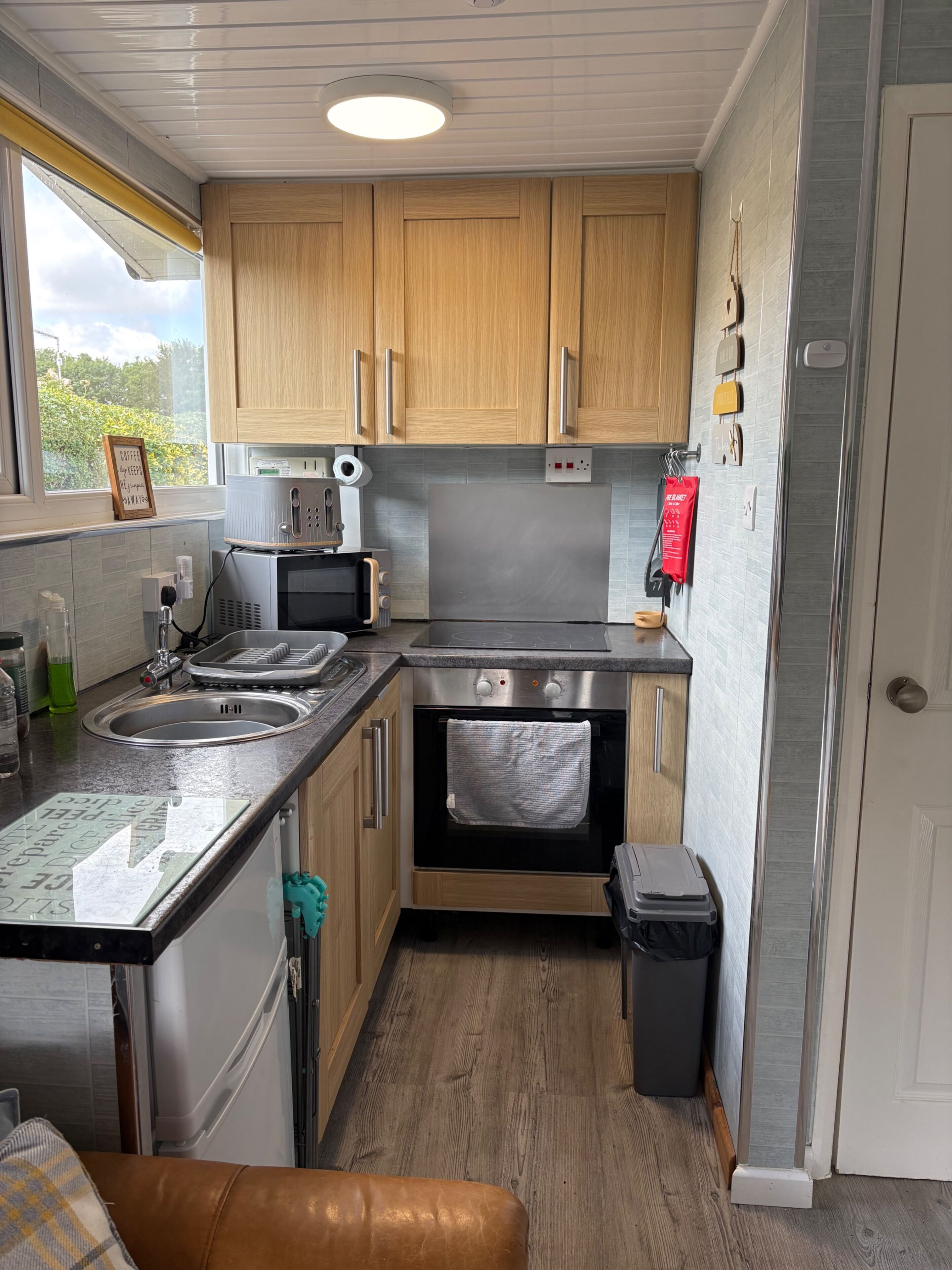 A compact kitchen with wooden cabinets, a stainless steel oven, a sink, and a small trash bin.