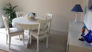 Et spiseområde med et rundt bord, hvide stole, en vase med blomster, en potteplante og en lampe.