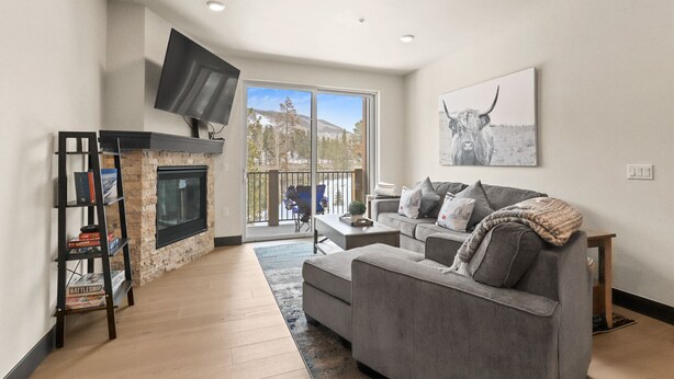 A living room with a fireplace, a sofa, a TV mounted on the wall, and a window with a view of trees and mountains. - Hot Tubs | Near Ski Resort & Restaurants (Keystone)