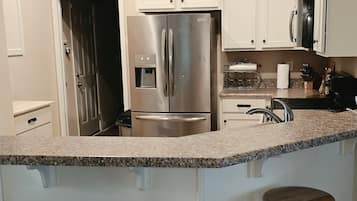 Una cocina con refrigerador de acero inoxidable, gabinetes blancos, encimera de granito y dos taburetes de madera.