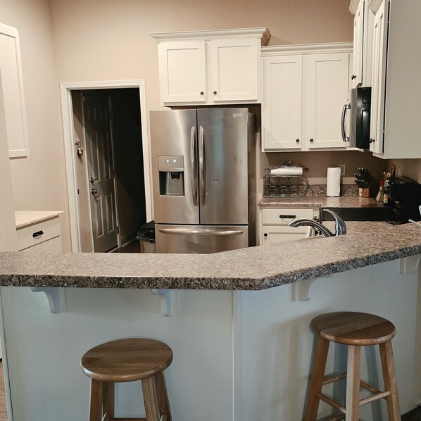 A kitchen with a stainless steel refrigerator, white cabinets, granite countertop, and two wooden stools.