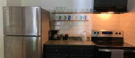 A kitchen with a stainless steel refrigerator, dark cabinets, a stove, and a shelf with glass cups.