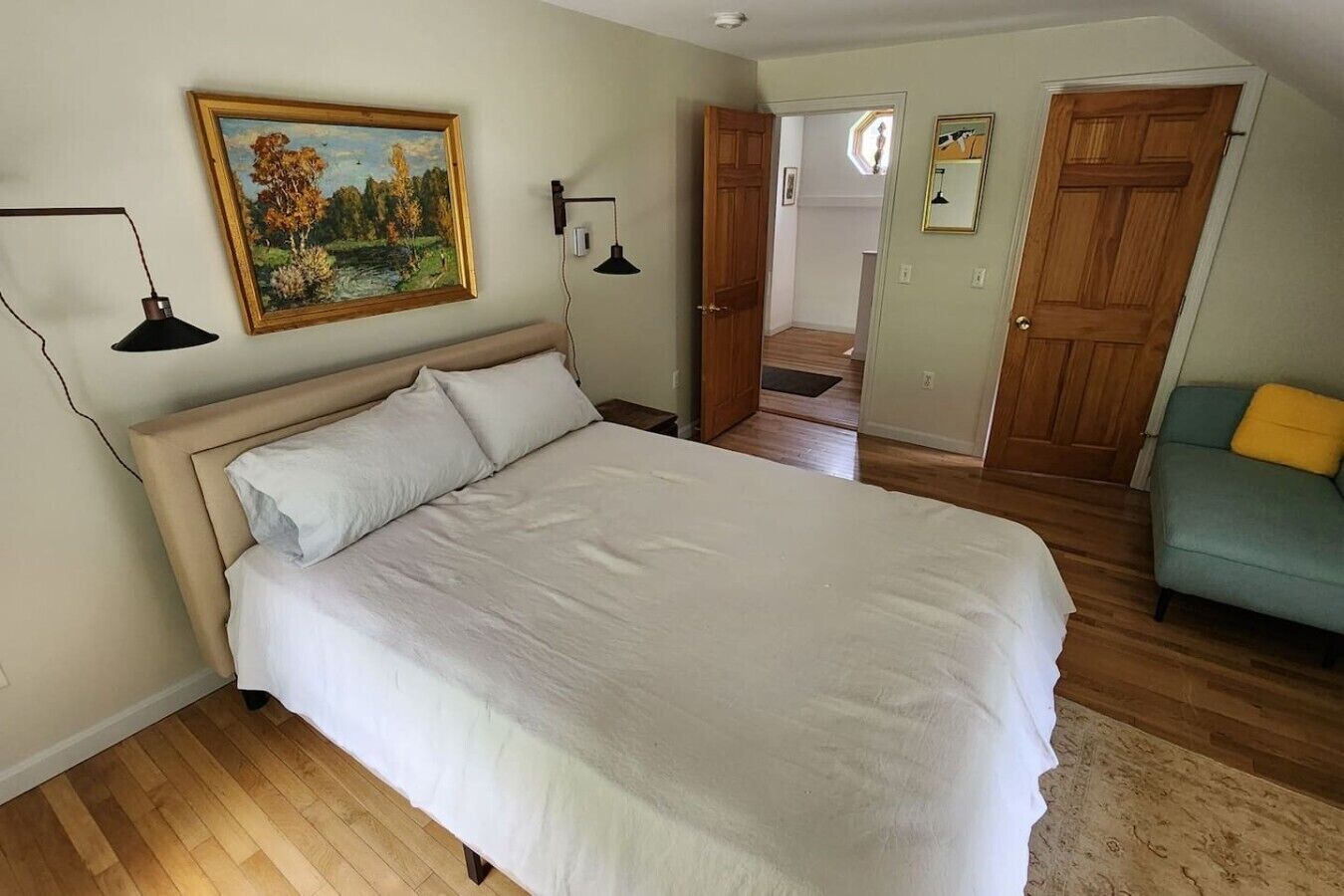 Une chambre à coucher avec un grand lit, un plancher en bois, un tableau accroché au mur et une porte donnant sur une autre pièce.