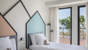 A hotel room with a bed, pillows, a teddy bear, a nightstand, a lamp, and a view of palm trees and a beach. - Finest Punta Cana (Punta Cana)