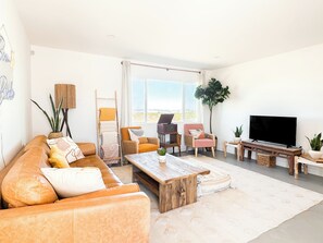 Ein Wohnzimmer mit einer Couch, Sesseln, einem Couchtisch, einem Fernseher und einem großen Fenster.