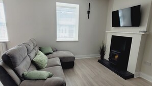 Ein Wohnzimmer mit Ecksofa, Kamin, einem an der Wand befestigten Fernseher und einem Fenster mit Jalousien.