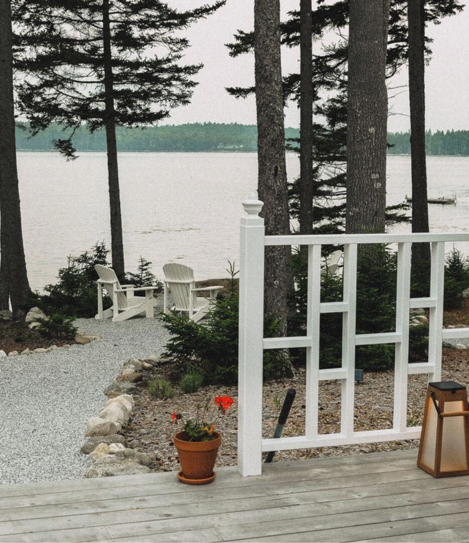 Um terraço com cadeiras Adirondack, cerca branca e vista para um lago.