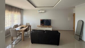 A modern living room with a dining table, a black sofa, a flat-screen TV, and an air conditioning unit on the wall.