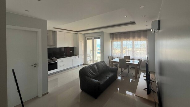 A modern living room with a black leather sofa, a dining area with a table and chairs, a kitchen with white cabinets and a black countertop, and a sliding glass door leading to a balcony.