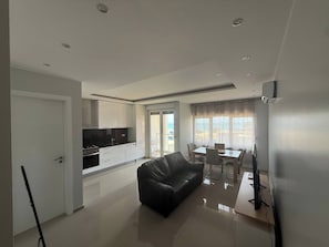 A modern living room with a black leather sofa, a dining area with a table and chairs, a kitchen with white cabinets and a black countertop, and a sliding glass door leading to a balcony.