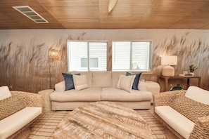 A living room with a beige sectional sofa, two windows with blinds, a lamp on a side table, and a woven rug. - Boutique Stay | Prvt Lounge, Arcade, Firepit | 15m IAH (Houston)