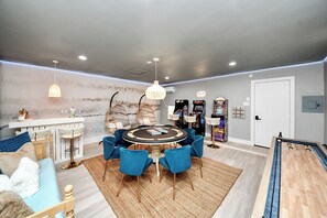 A room with a round table, blue chairs, a bar area, arcade games, and a foosball table. - Boutique Stay | Prvt Lounge, Arcade, Firepit | 15m IAH (Houston)