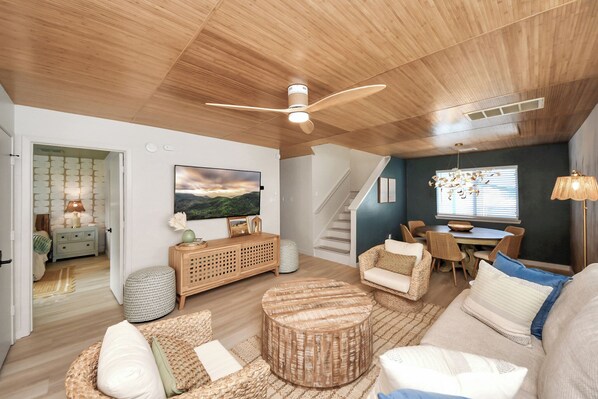 A spacious living room with a wooden ceiling, a large flat-screen TV, a dining area with wicker furniture, and a staircase leading upstairs.