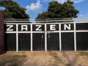 A gated entrance with the word 'ZAZEN' painted on it, surrounded by a white fence and trees in the background.