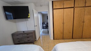 Une chambre avec deux lits, un téléviseur fixé au mur, une armoire en bois et un couloir menant à un espace de vie.